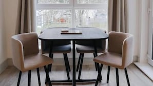Et spiseområde med et rundt bord og to stole, et vindue med gardiner, og udsigt til træer udefra.