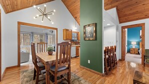 A spacious dining area with a wooden table and chairs, a ceiling light fixture, and a view of the kitchen in the background.