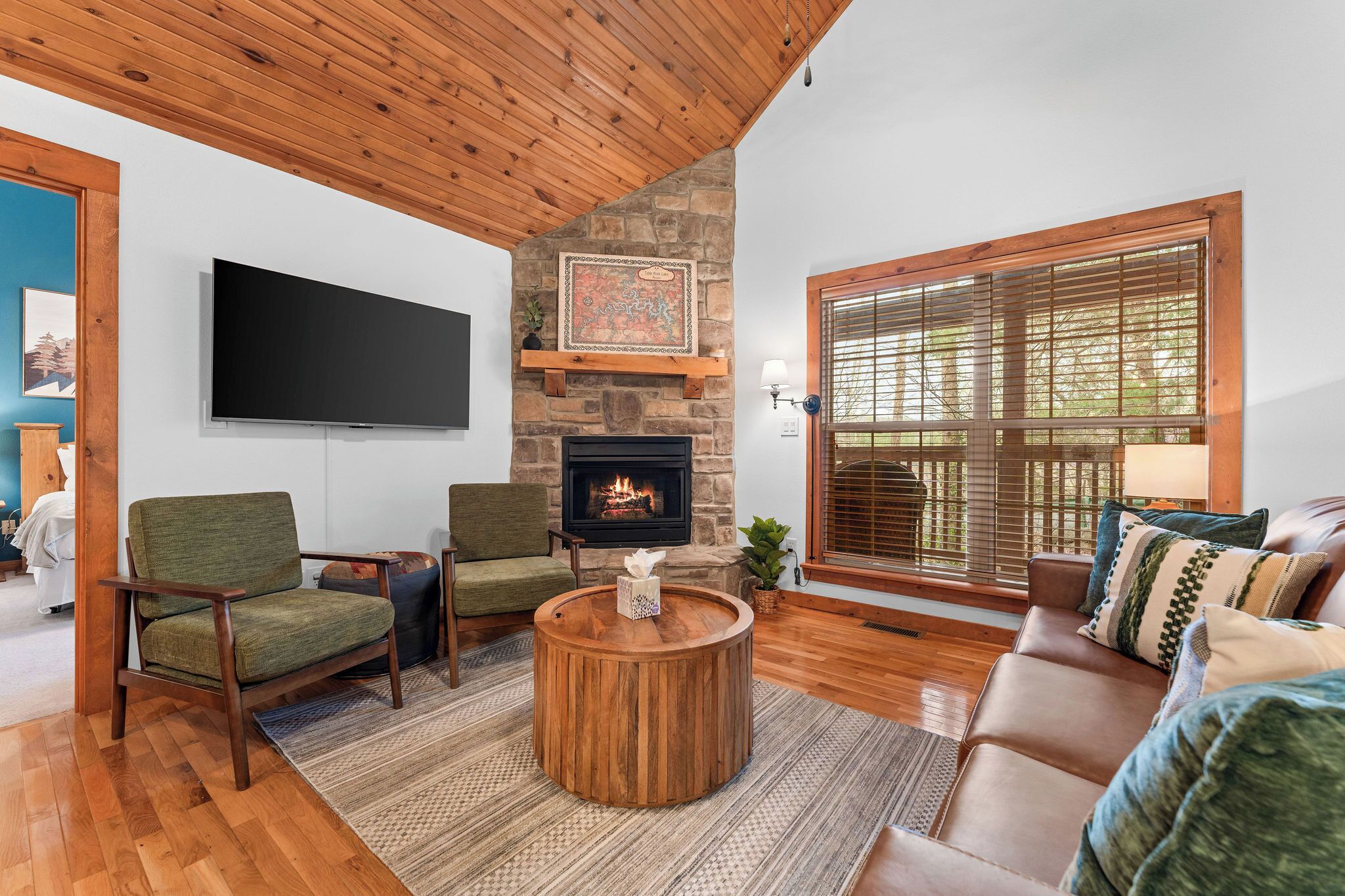 A cozy living room with a stone fireplace, wooden ceiling, and a large window with blinds.