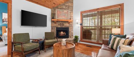 A cozy living room with a stone fireplace, wooden ceiling, and a large window with blinds.