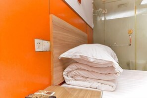 A hotel room with an orange wall, a wooden headboard, a white bed with a folded white comforter, a bedside table with a remote control, and a glass shower door. - Haoting hotel (Shanghai Xincun Road Subway Station) (Shanghai)