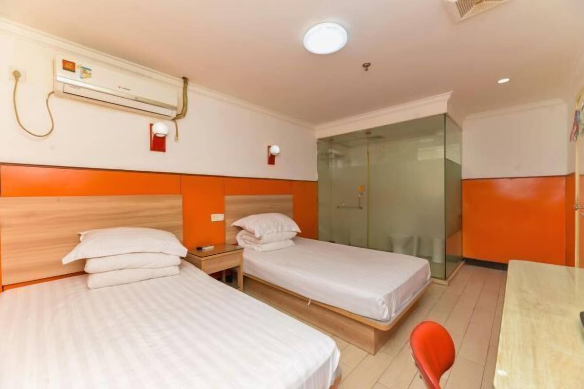 A hotel room with two beds, a desk, a chair, an air conditioning unit, and a glass shower enclosure.