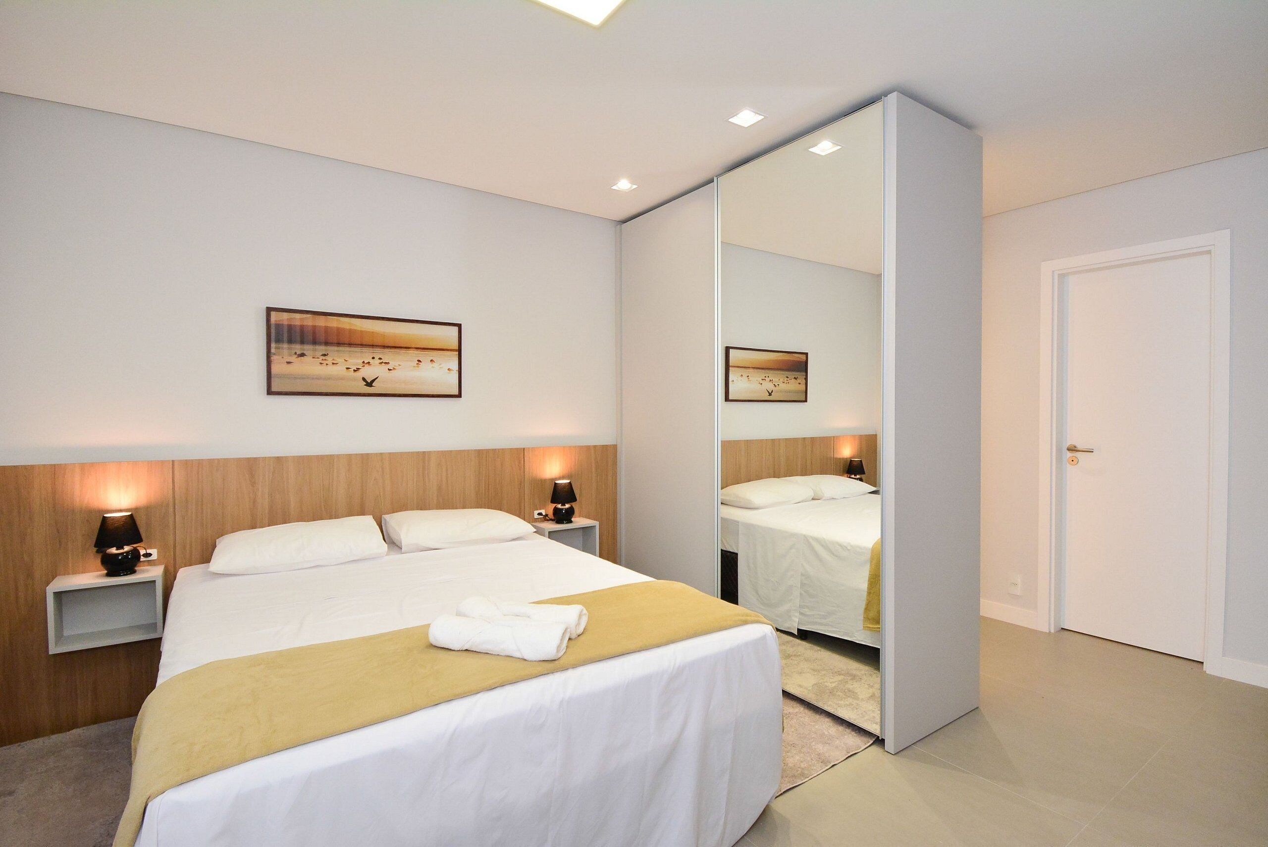 A modern hotel room with a large bed, bedside tables with lamps, a mirror, and a painting on the wall.