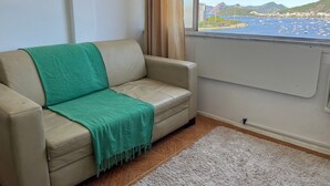 Un canapé beige agrémenté d’un plaid vert, un tapis blanc et une vue sur une ville côtière avec des bateaux et des montagnes.