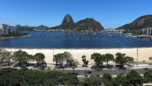 Un paysage urbain côtier avec une montagne imposante, une marina remplie de bateaux et une plage bordée d’arbres et d’une route.