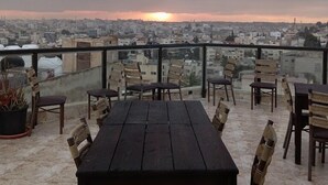 Terraço no telhado com mesa e cadeiras de madeira, com vista para a paisagem urbana ao pôr do sol.