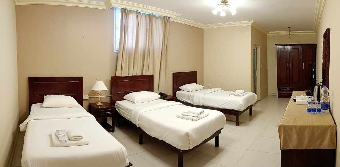 A hotel room with three single beds, a bedside table with a lamp, a desk with a chair, and a wardrobe.