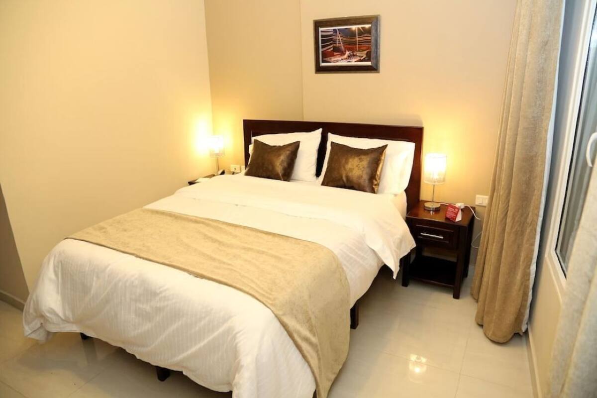 A neatly made bed with a beige bedspread and pillows, a bedside table with a lamp, and a framed picture on the wall.