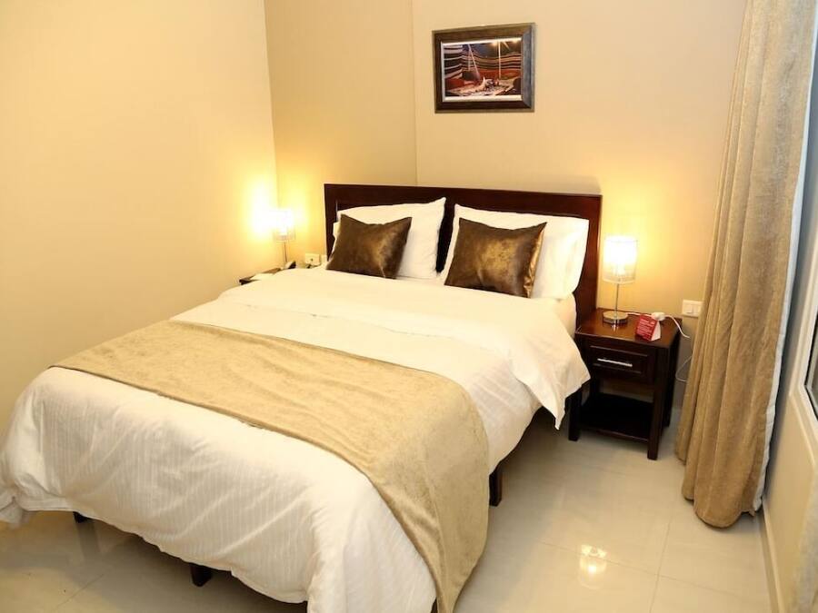 A neatly made bed with a beige bedspread and pillows, a bedside table with a lamp, and a framed picture on the wall.