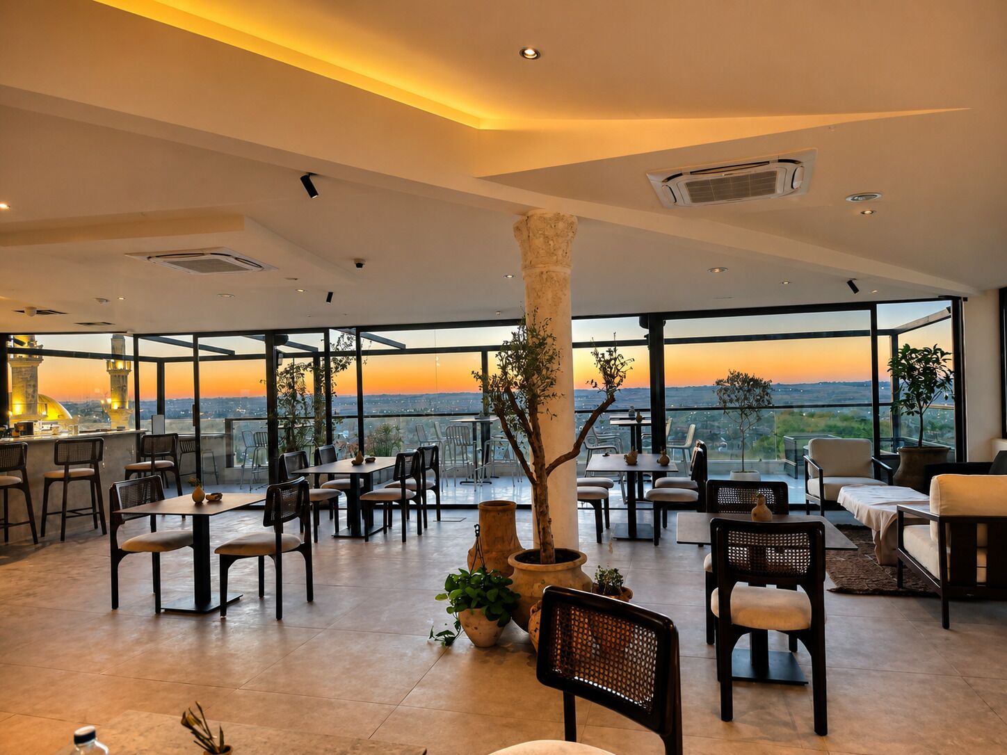 Un comedor amplio con varias mesas y sillas, ventanas grandes que ofrecen vista al atardecer y un column central con un árbol.