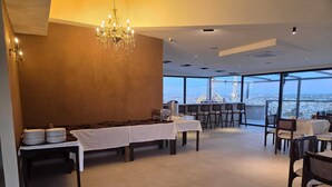 Uma sala de jantar espaçosa, com pé direito alto, um candelabro e vista para a cidade através de grandes janelas.