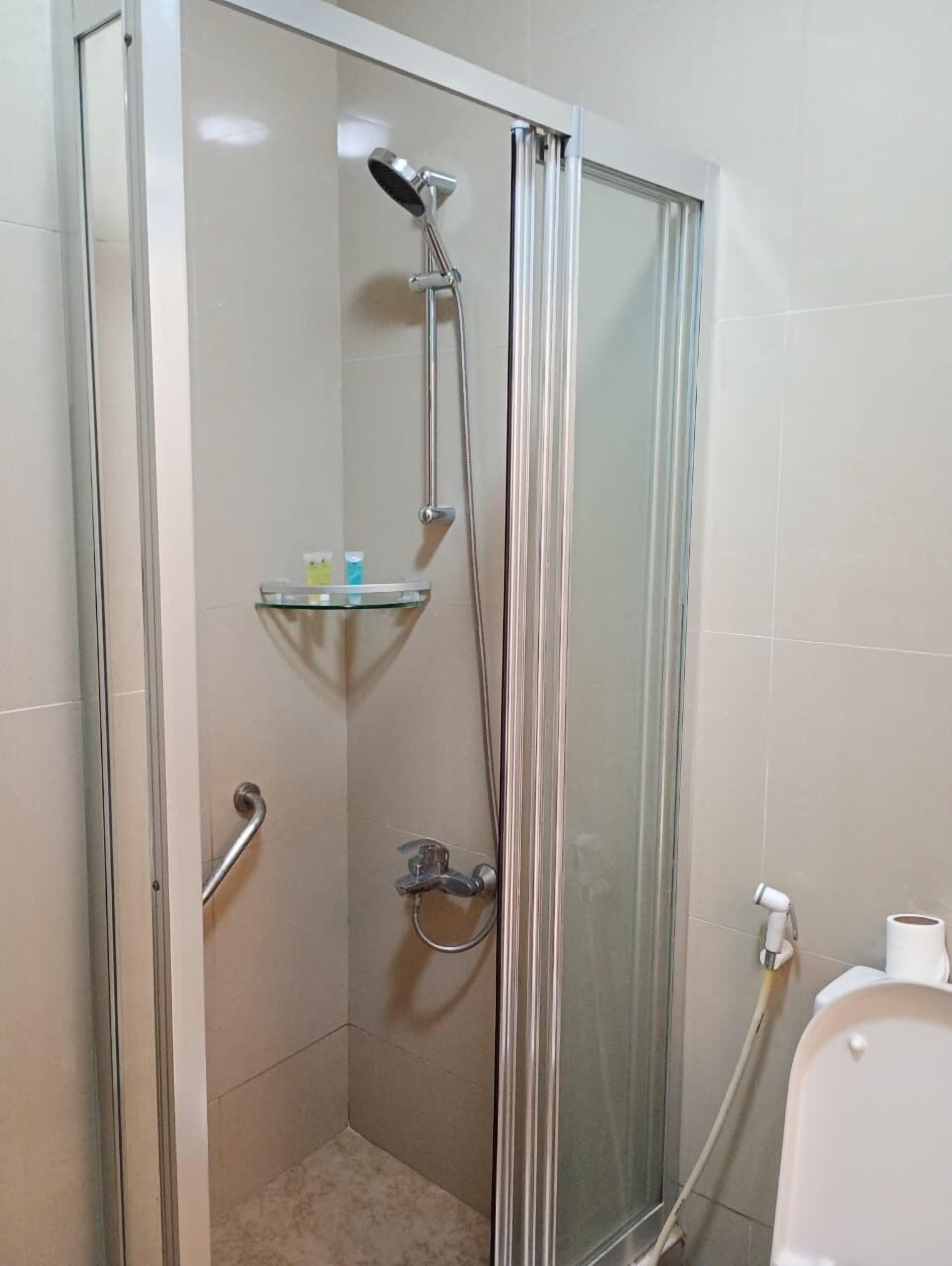 A glass-enclosed shower stall with a handheld showerhead, a handheld shower attachment, and a glass shelf with toiletries.
