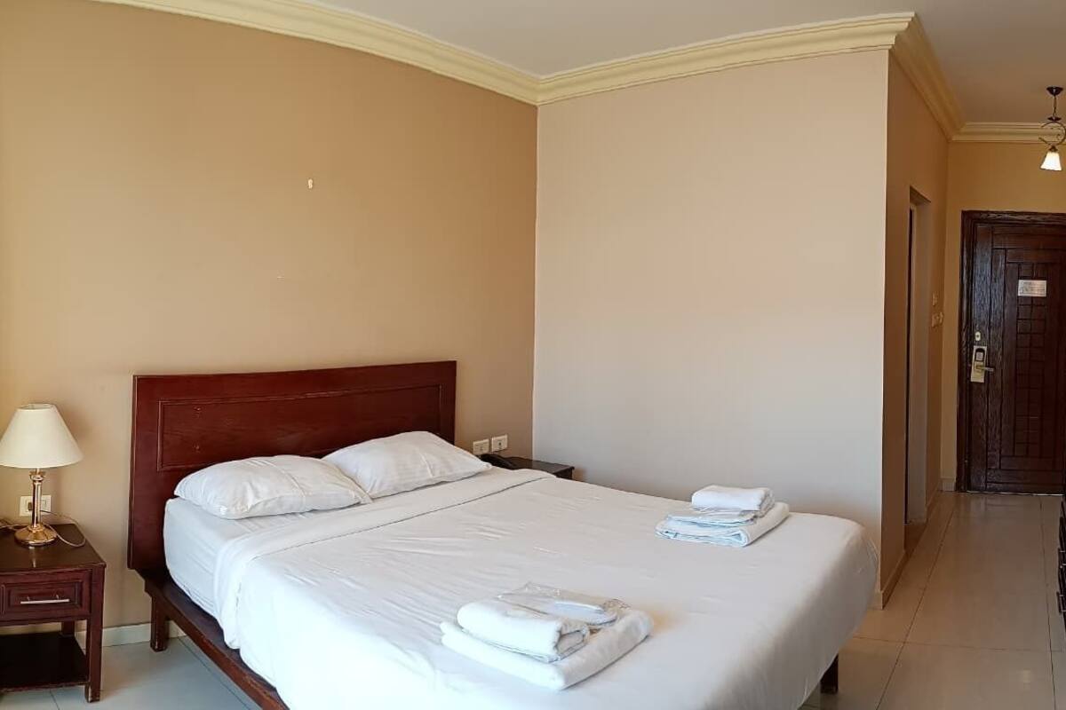 A hotel room with a large bed, a nightstand with a lamp, and a door leading to another room.
