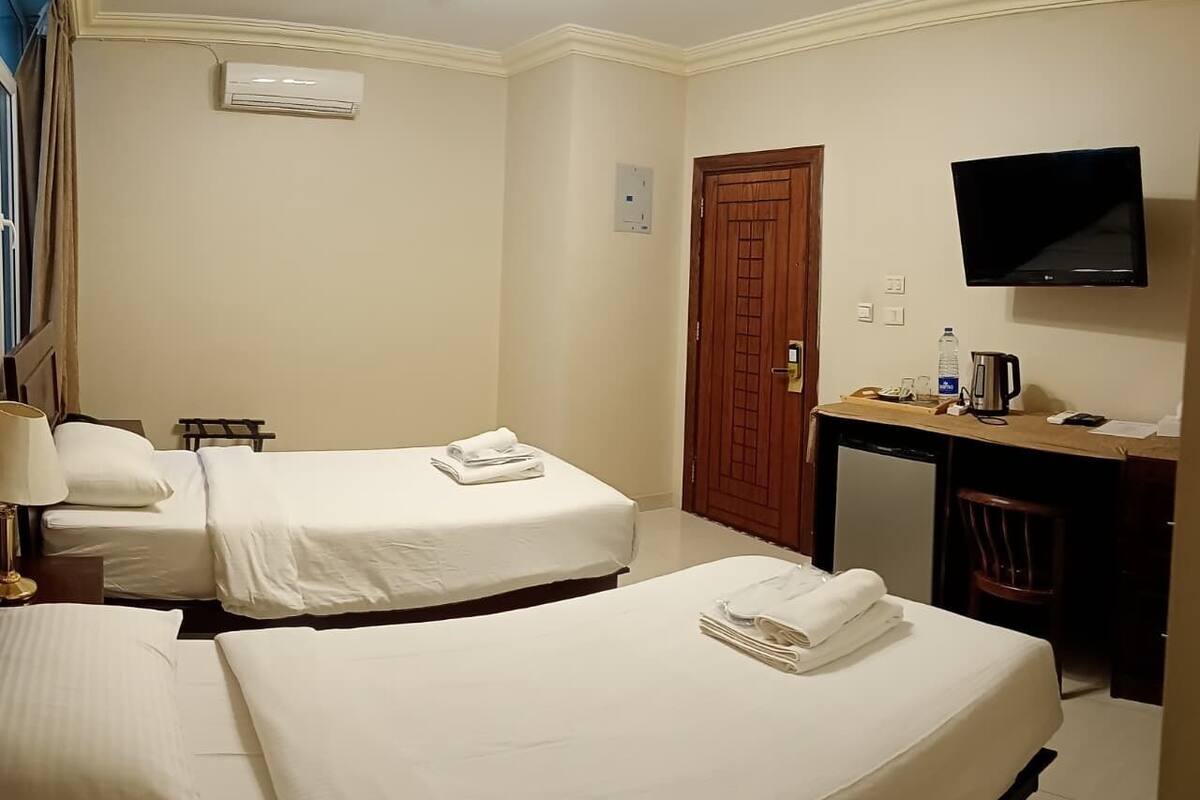 A hotel room with two beds, a desk with a chair, a television, and an air conditioning unit.