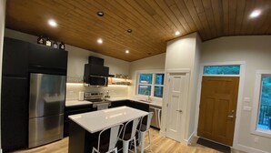 A modern kitchen with a central island, bar stools, a stainless steel refrigerator, and a wooden ceiling.