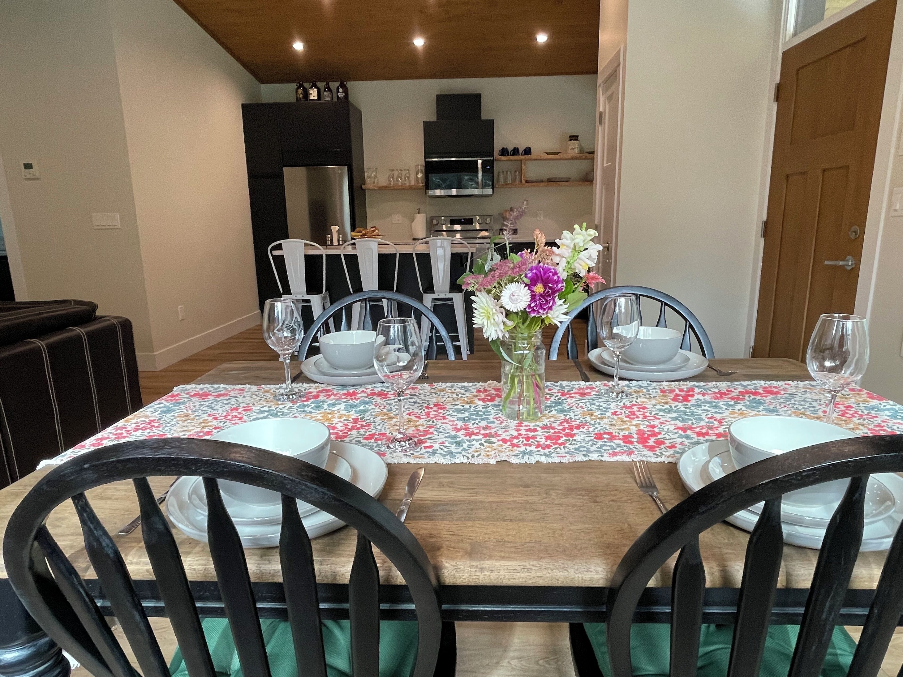 A dining area with a wooden table set for four, surrounded by black chairs. A vase with flowers is placed in the center.