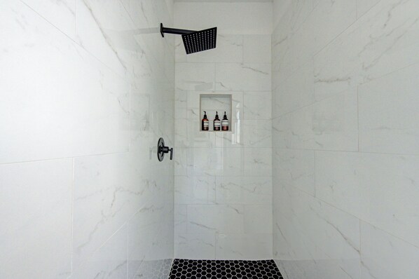Une douche équipée d’un pommeau de douche noir à main, d’une petite étagère avec des bouteilles et d’un drain hexagonal.