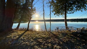 Une scène au bord d’un lac, avec un banc et une table de pique-nique sous les arbres, un ciel dégagé et le soleil se reflétant sur l’eau.