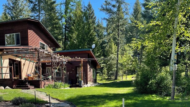A wooden cabin with a stone pathway leading to the entrance, surrounded by a lush green landscape and tall trees. - Charming 1-bedroom apartment in a beautiful natural setting, minutes from town. (Sandpoint)