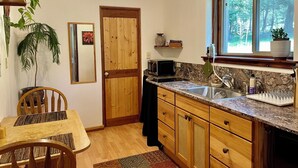 A compact kitchen with wooden cabinets, a granite countertop, a sink, and a window with a view of trees. - Charming 1-bedroom apartment in a beautiful natural setting, minutes from town. (Sandpoint)