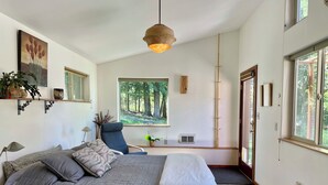 A bedroom with a large bed, a desk, a chair, a window with a forest view, and a hanging light fixture. - Charming 1-bedroom apartment in a beautiful natural setting, minutes from town. (Sandpoint)