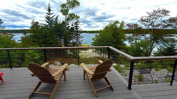 A deck with two wooden chairs and a table, overlooking a lake with trees and a cloudy sky.