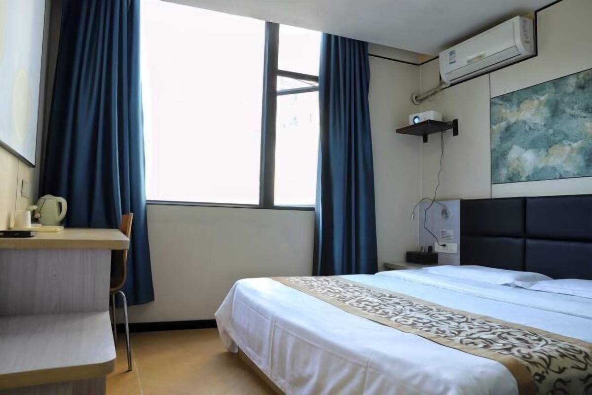 A hotel room with a bed, a desk, a chair, a window with curtains, an air conditioner, and a wall-mounted shelf.