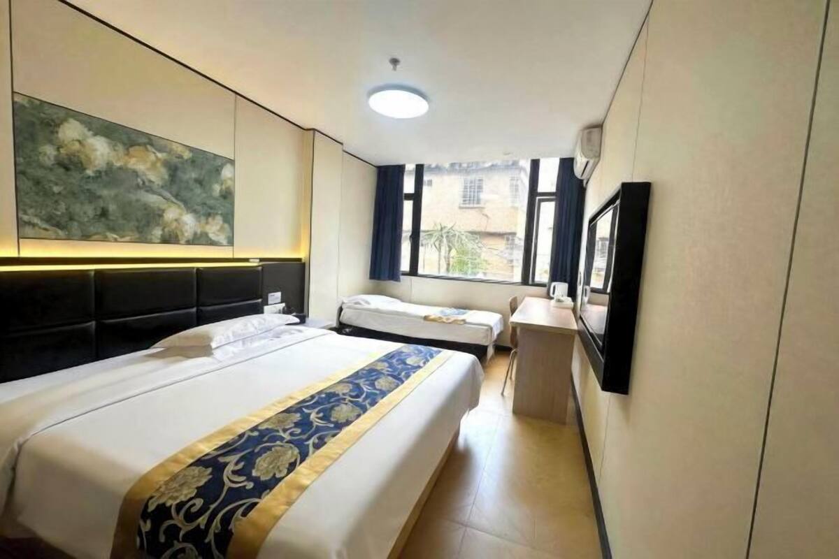 A hotel room with a large bed, a smaller bed, a desk, a chair, a window, and a wall-mounted TV.