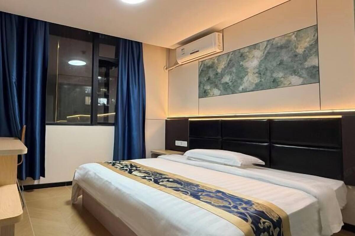 A hotel room with a bed, a desk, a window with curtains, an air conditioning unit, and a wall-mounted artwork.
