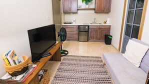 Un espace de vie compact comprenant un téléviseur à écran plat, un ventilateur, un canapé, une machine à café et un petit coin cuisine avec des placards et un évier.