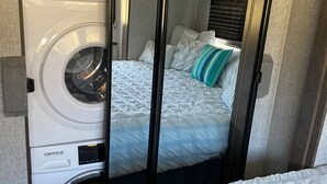 A compact room with a washing machine, a bed with a quilted cover, and a window with blinds. - Charming 2-bedroom RV with WiFi and AC in peaceful Silverhill Alabama (Silverhill)