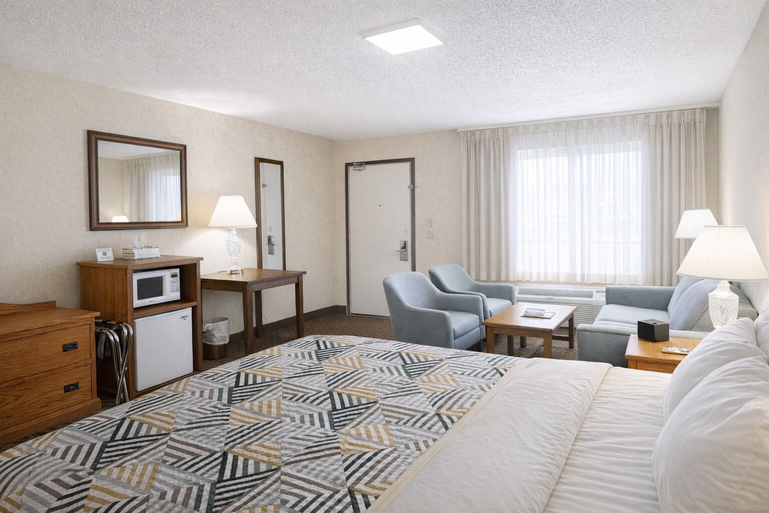 A hotel room with a bed, a desk with a microwave, two armchairs, a sofa, and a large window with curtains.