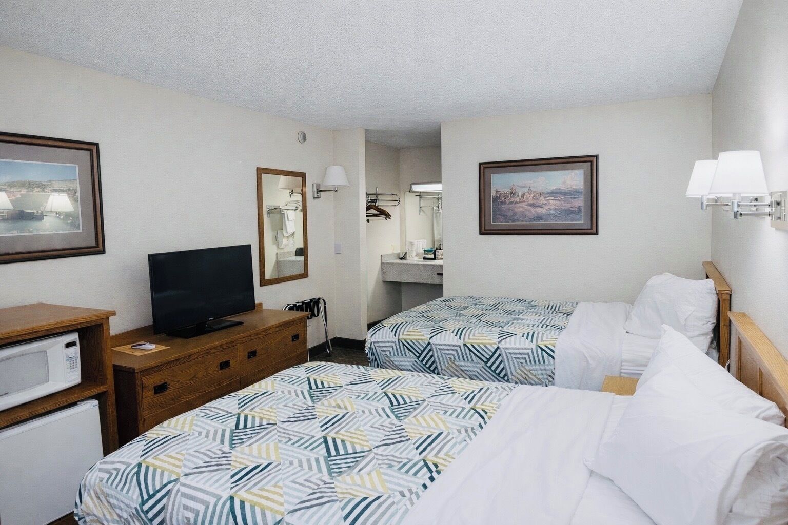 Ein Hotelzimmer mit zwei Betten, einem Fernseher, einem Schreibtisch mit Mikrowelle und Bildern an den Wänden.