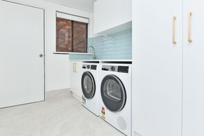 A laundry room with two front-loading washing machines, a window with a view of a brick wall, and a white door. - Luna at Corlette Beach House with views (Corlette)