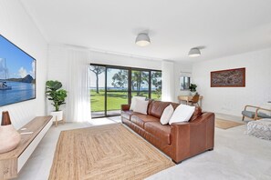 A spacious living room with a large window, a brown leather sofa, a wooden coffee table, and a view of the sea.