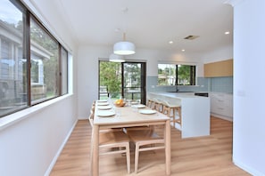 A modern kitchen with a central island, large windows, and a dining area with a table set for six. - Luna at Corlette Beach House with views (Corlette)