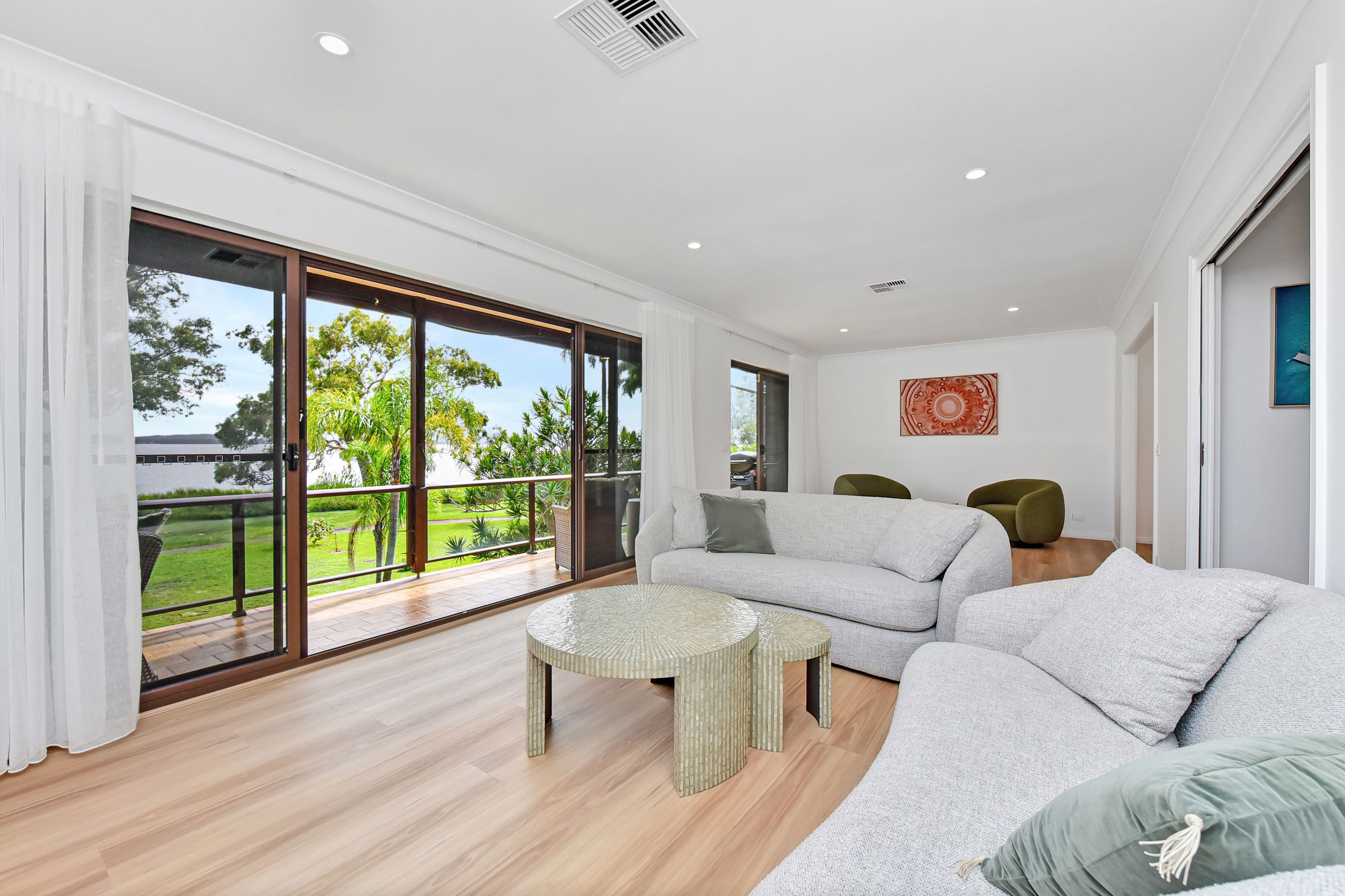 A spacious living room with a large sliding glass door leading to a balcony, a grey sofa set, a round coffee table, and a wall-mounted artwork.