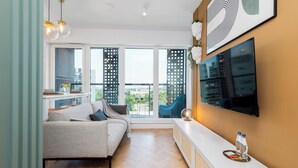 A modern living room with a sofa, a TV mounted on the wall, and a balcony with a view of buildings. - Apartament Tower by Kairos Apartments (Katowice)