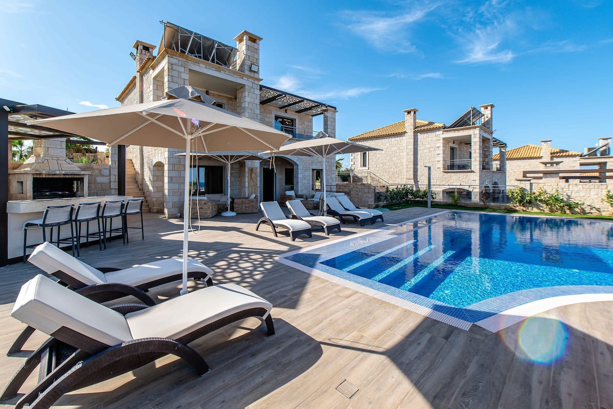 Un'area piscina con sedie a sdraio e ombrelloni, un edificio in pietra con camino e un cielo azzurro limpido.
