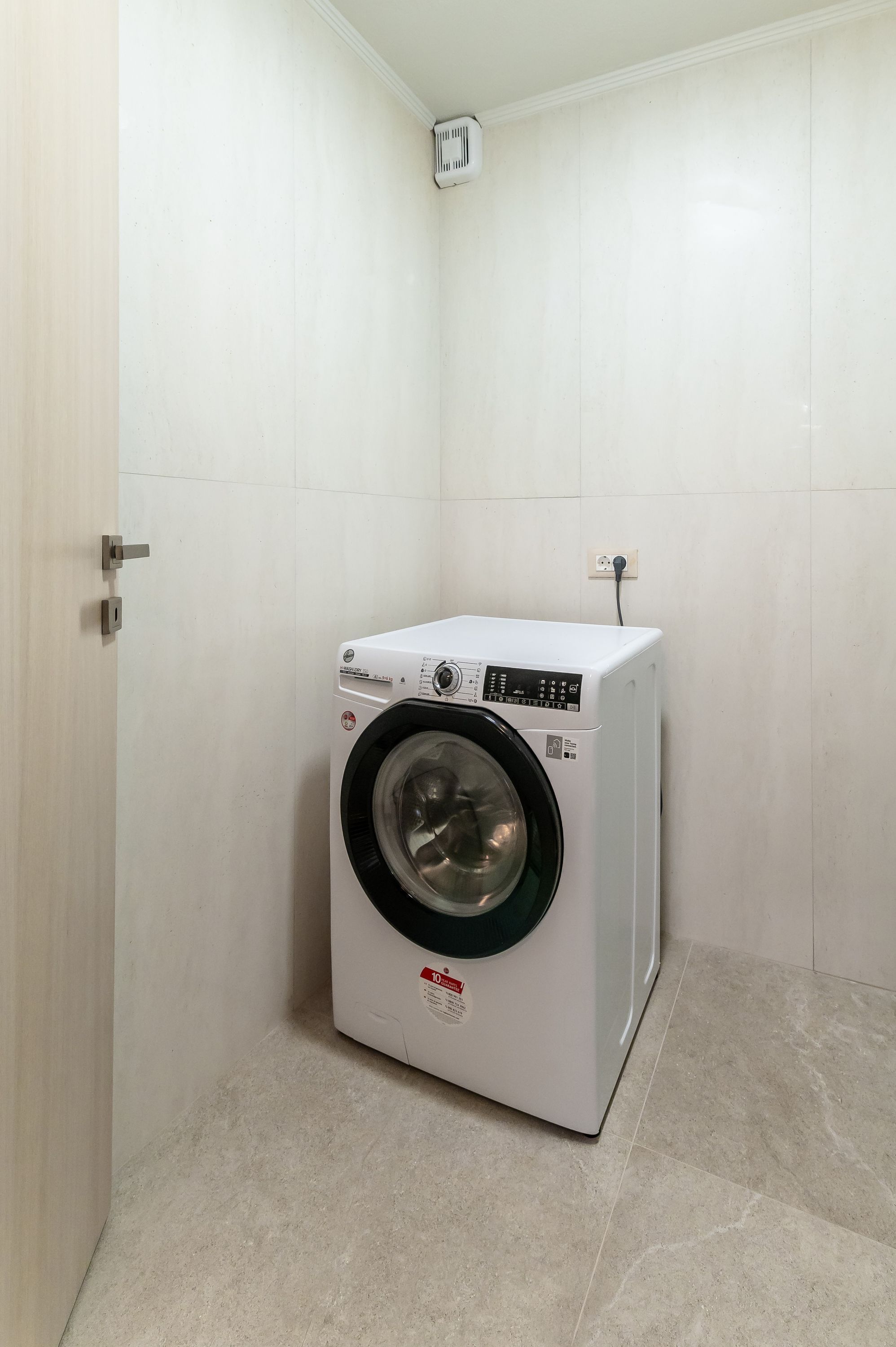 A compact, white washing machine in a small, tiled bathroom with a door and a power outlet on the wall.