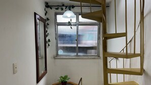 Una habitación con escalera de caracol, una ventana con persianas, un cuadro en la pared y una pequeña mesa octogonal con una planta.