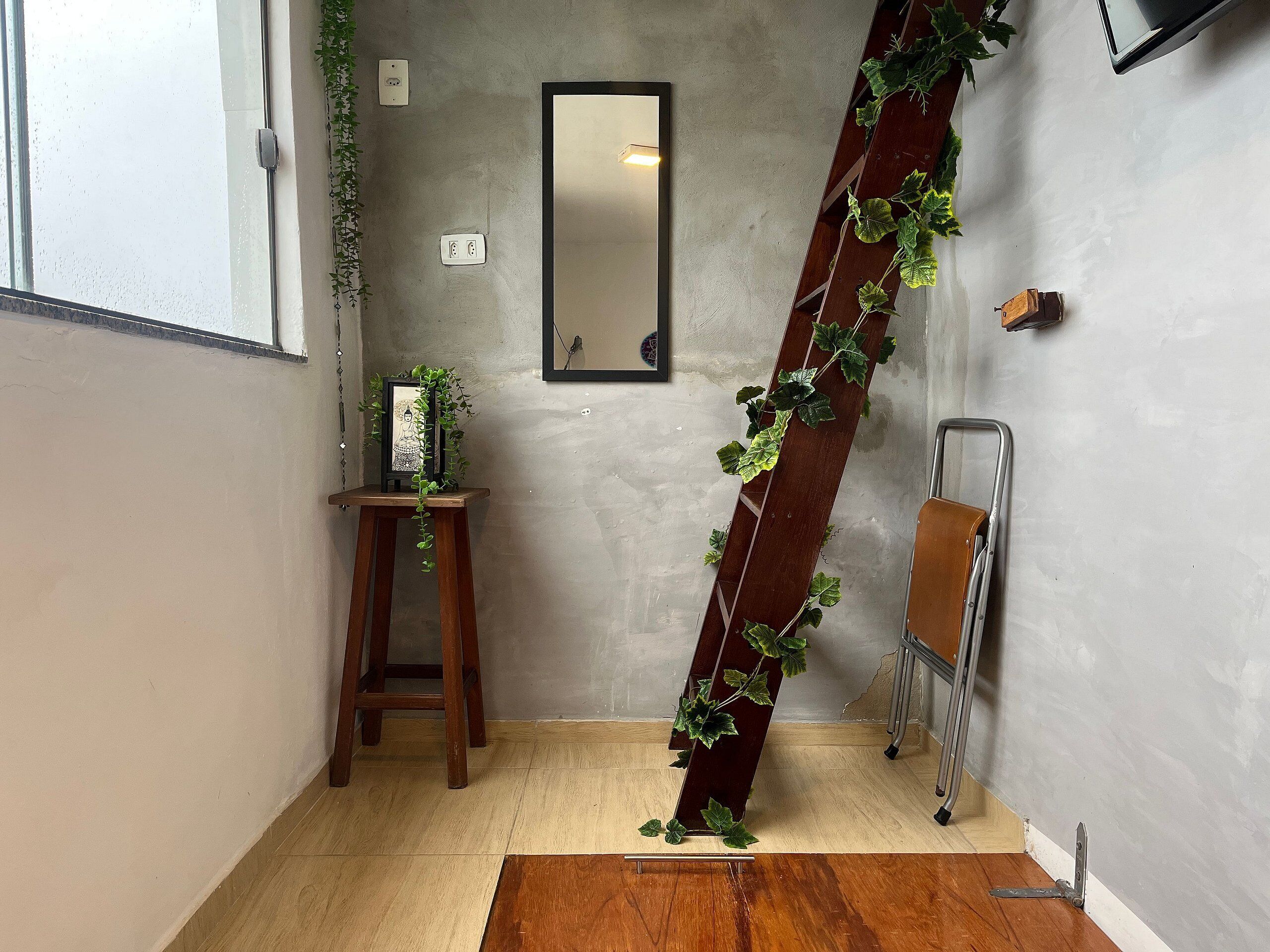 Una habitación con piso de madera, un taburete de madera pequeño, un espejo en la pared y una planta sobre el taburete.