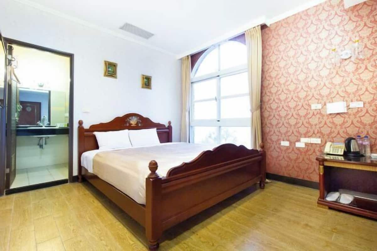 Un dormitorio con una cama de madera, un vidrio que da a un baño, un ventanal grande y un papel tapiz decorado.