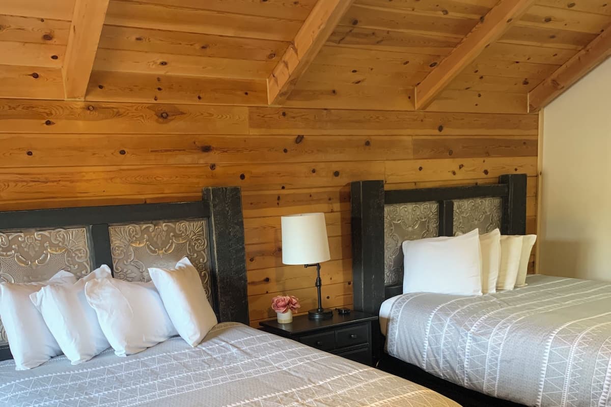 A room with two beds, wooden ceiling beams, a nightstand with a lamp, and a wall with a decorative headboard.
