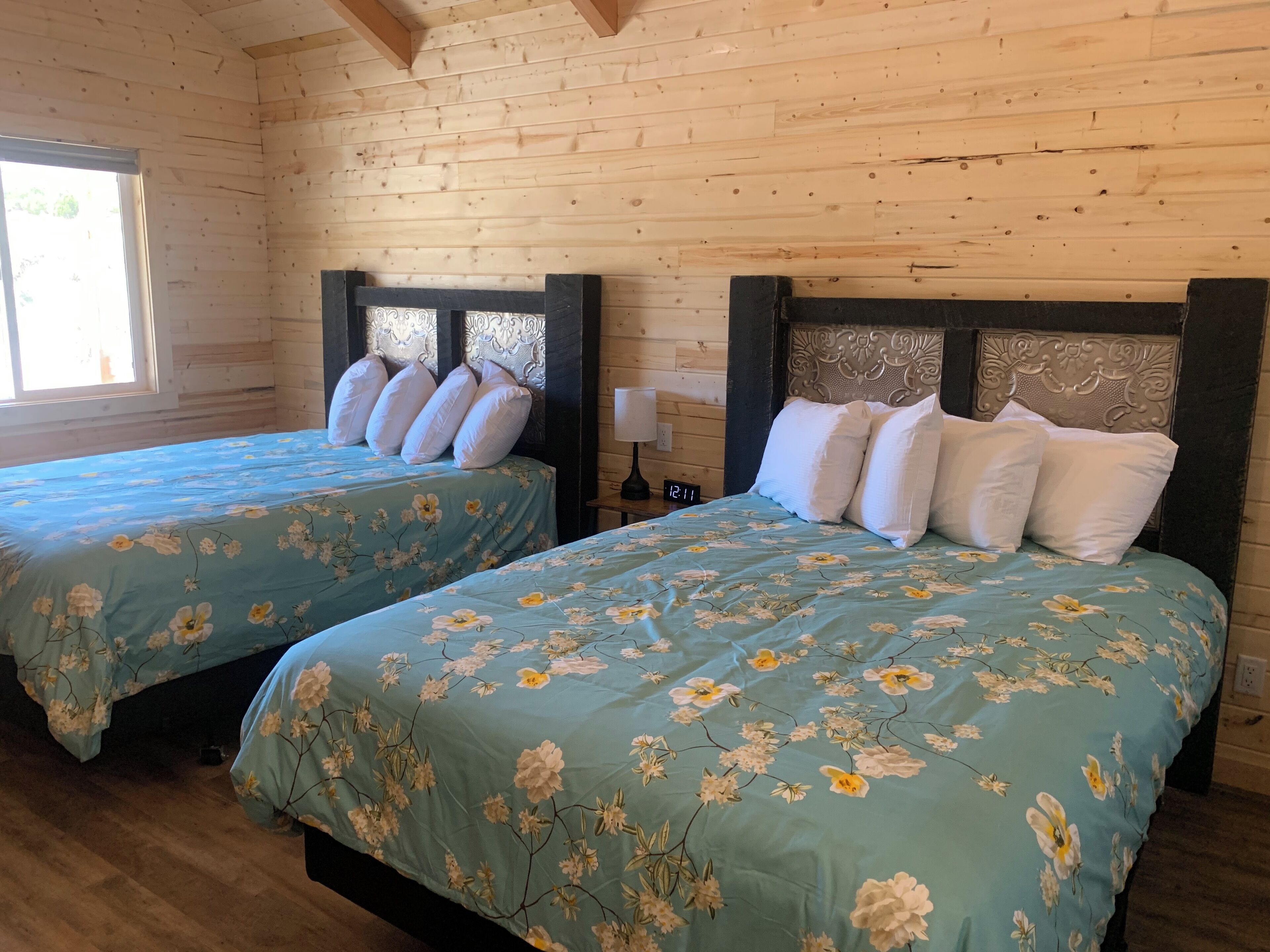 A room with two beds, each with a floral bedspread and matching pillows, wooden walls, and a window with daylight coming through.