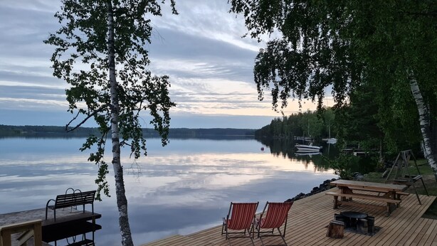 Una terraza de madera con bancos y sillas da a un lago tranquilo, rodeado de árboles.