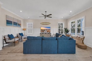 A spacious living room with a large blue sectional sofa, a ceiling fan, and a view of the outdoors through large windows.