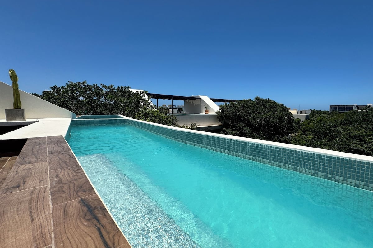 A modern rooftop pool with clear blue water, surrounded by lush greenery and a clear sky.
