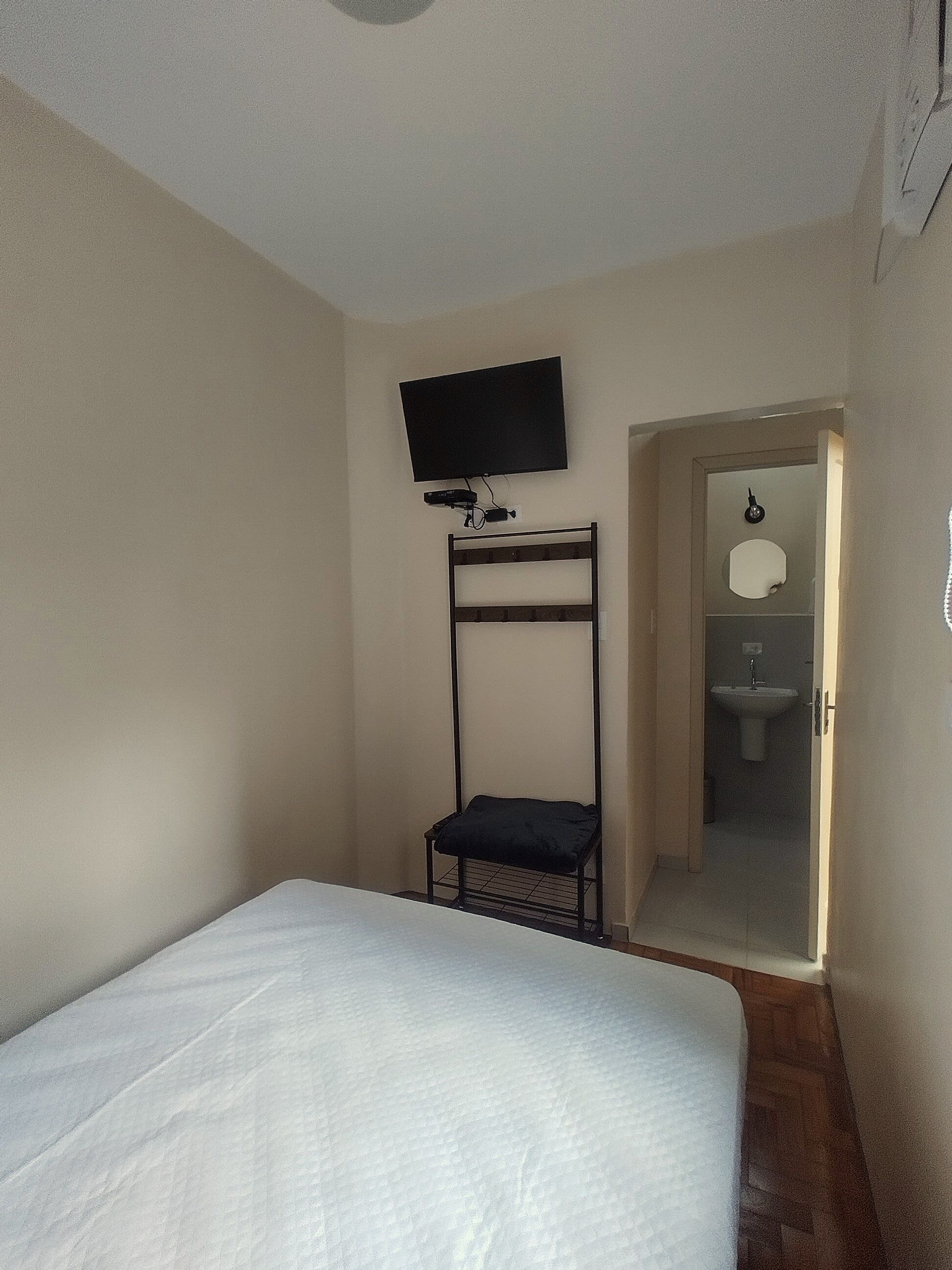 Une chambre à coucher comprenant un lit, un téléviseur fixé au mur, une chaise et une porte donnant sur une salle de bain.