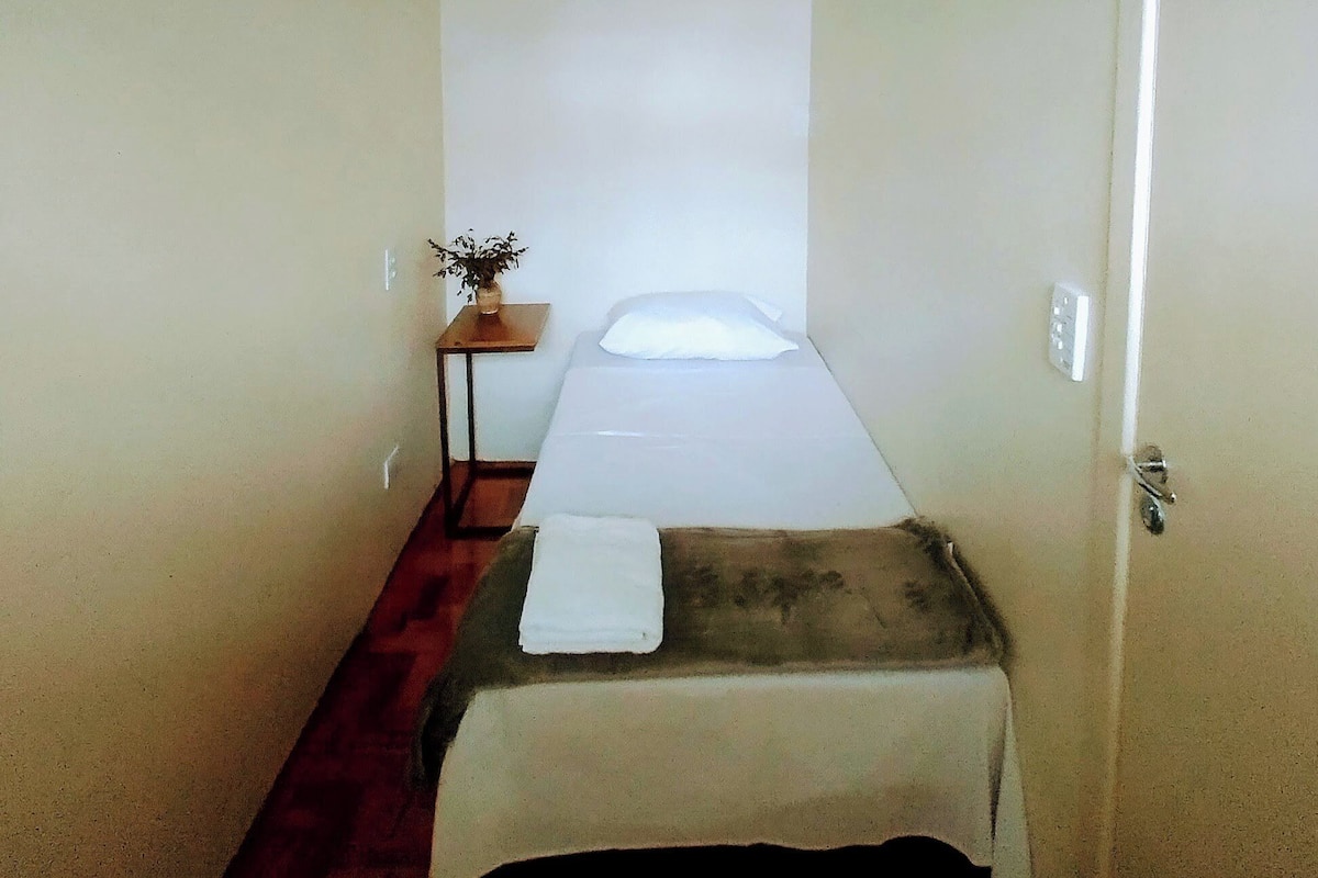 Ein schmales Hotelzimmer mit einem Einzelbett, einem kleinen Tisch mit Pflanze und einer Tür rechts.